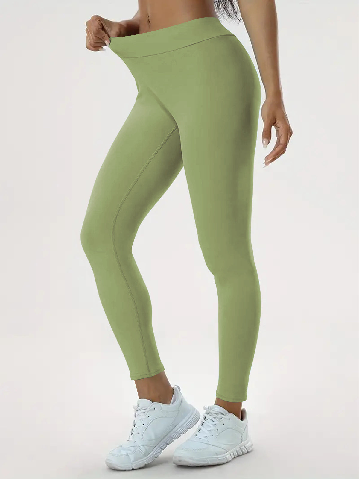 "Person wearing light green leggings and white athletic shoes, demonstrating the stretchiness and fit of the leggings by pulling the waistband away from their body."