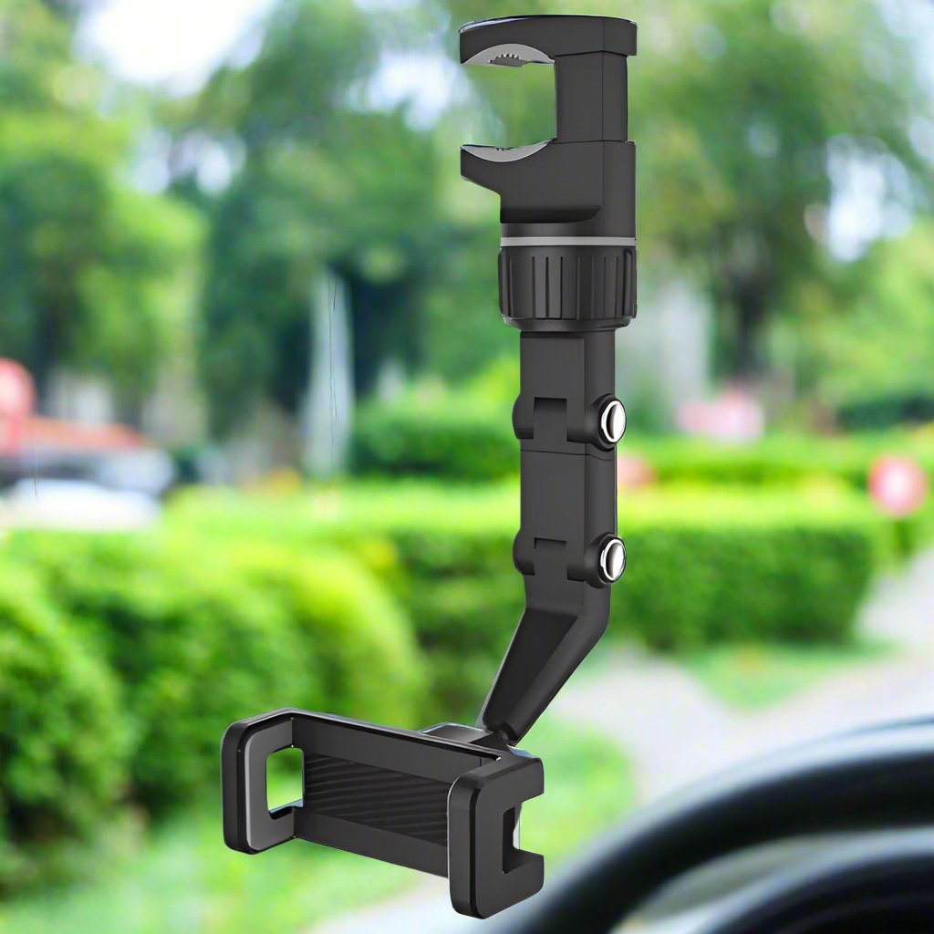 An adjustable car phone mount holder with a sturdy, black design featuring a clamp mechanism. The mount has a rotating and extendable arm for optimal phone positioning. The background shows a blurred outdoor scene with greenery, indicating the mount's use in a car setting.