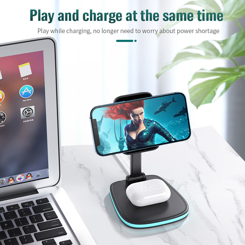 "A smartphone and wireless earbud charging station on a marble surface. The station holds a smartphone in landscape orientation displaying an animated character and has a pair of wireless earbuds charging below. A laptop is partially visible on the left side of the image. Text at the top reads 'Play and charge at the same time. Play while charging, no longer need to worry about power shortage.'"