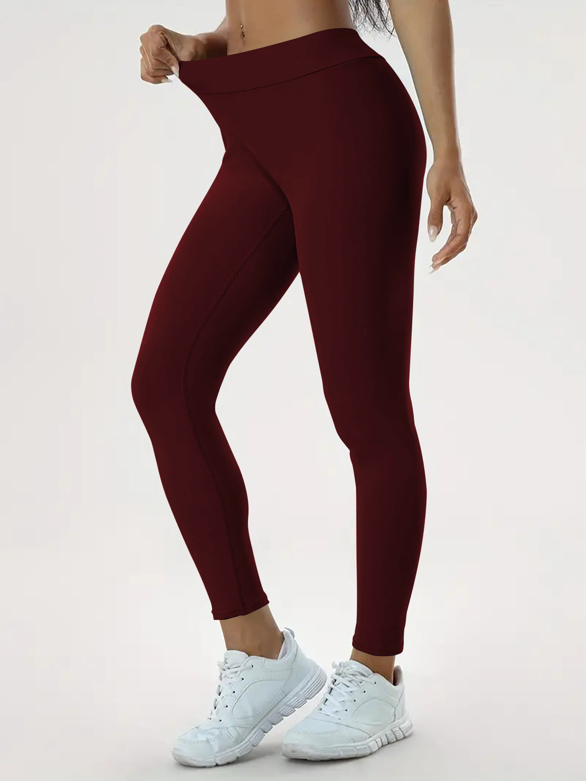  A person wearing burgundy high-waisted stretchy leggings and white sneakers, pulling the waistband to demonstrate the fit and flexibility against a light-colored background.

