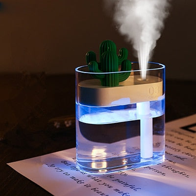 "Alt text: A small, transparent humidifier with a cactus-shaped decoration on top emits a stream of mist. The humidifier is illuminated with a blue light and is placed on a piece of paper with text."