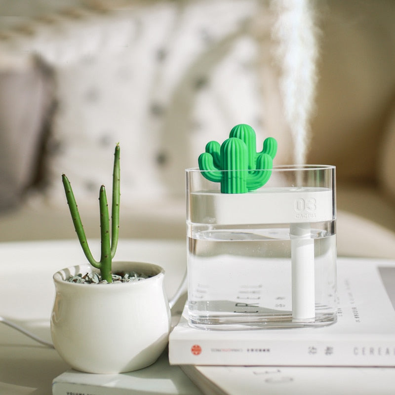 "A small green cactus-shaped humidifier is placed on a white table next to a small potted plant in a white pot. The humidifier is emitting a fine mist. The table also has a stack of books, with one book partially visible and titled 'Cereal'."