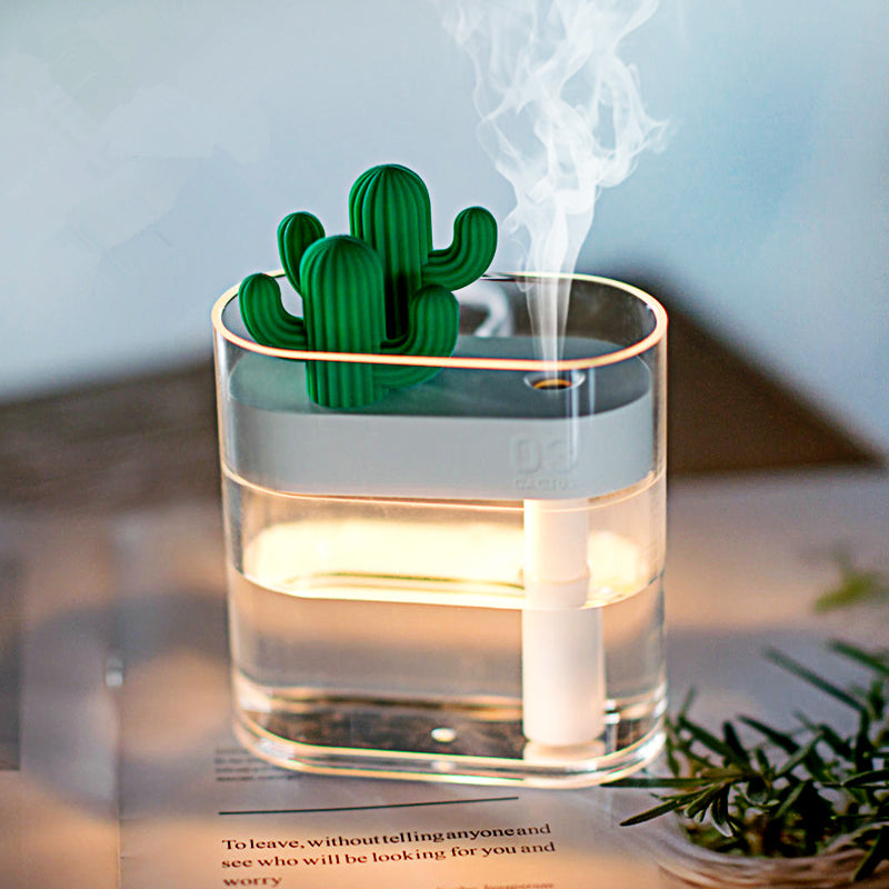 "A small, transparent humidifier with a green cactus-shaped decoration on top emits a stream of mist. The humidifier is placed on a surface with a blurred background that includes a book or magazine and some greenery."