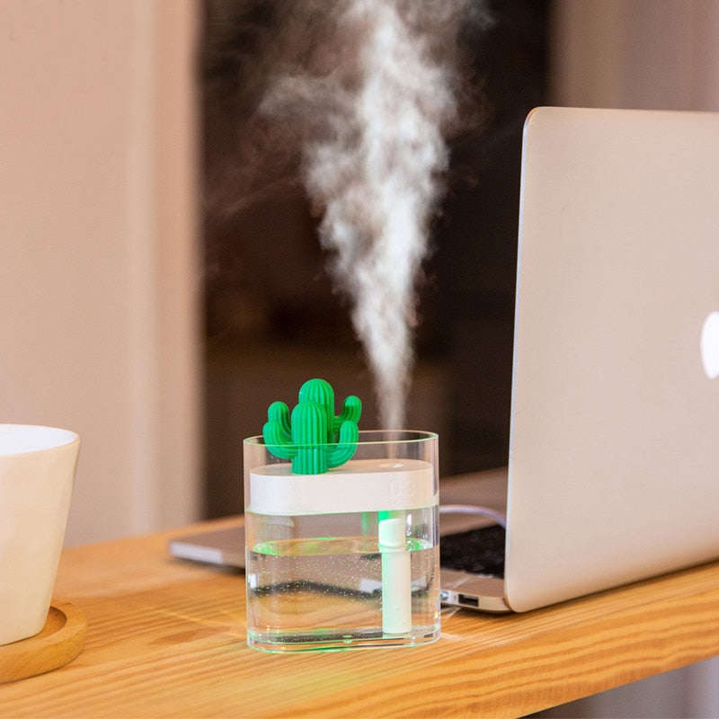 "A small cactus-shaped humidifier is placed in a transparent glass of water on a wooden table. The humidifier is emitting a stream of mist. A laptop is positioned nearby on the table, and part of a white cup on a wooden coaster is visible on the left side of the image."